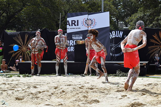 ive performers in red cloth and white body paint perform a traditional dance on a sandy surface during an outdoor cultural festival. A backdrop with the words "Yabun" and logos is visible, with audience members seated on grass and trees lining the background.