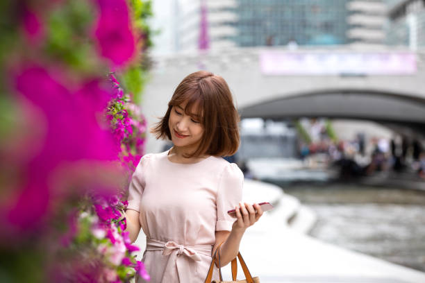 A woman in a light pink dress holding a brown handbag and a smartphone, standing near a vertical garden of pink flowers with a stone bridge and waterway in the background."
